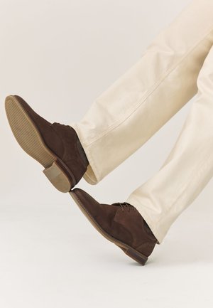 Jambes en pantalon beige portant des chaussures habillées à lacets en daim marron foncé, un pied levé montrant le détail de la semelle et de la bande de roulement du talon.
