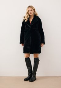 Zwarte nepbontjas met grote kraag, open voorkant en lange mouwen, gedragen over een rode top. Gecombineerd met zwarte kniehoge laarzen.