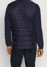 Mörkblå hybridjacka med ett quiltat, vadderat bakstycke, långa åtsittande ärmar, slim fit och elastiska mudd. Slät tyg med en subtil lyster.