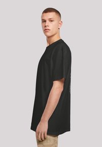 Black cotton T-shirt with a relaxed fit, short sleeves, and a rounded neckline, featuring a graphic design on the back. Model posed sideways.