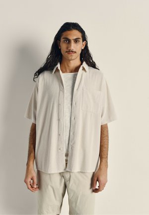 Chemise courte à manches courtes beige clair avec une texture côtelée verticale et une poche sur la poitrine, portée par-dessus une chemise gris clair et assortie à un short beige.