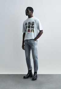 Άνδρας φορώντας γυαλιά ηλίου, λευκό t-shirt με τυπωμένα σχέδια καπέλων, ξεθωριασμένο γκρι τζιν και μαύρα δερμάτινα μποτάκια, που στέκεται μπροστά σε απλό φόντο.