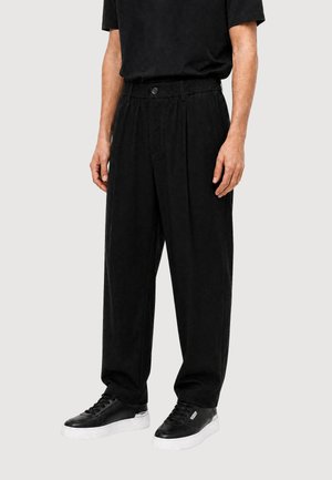 Uomo che indossa pantaloni neri larghi, t-shirt nera e scarpe da ginnastica nere con suole bianche, in piedi davanti a uno sfondo chiaro e semplice.