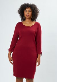 Rote Strickkleid mit einem wellenförmigen Ausschnitt und dreiviertel Ärmeln. Verfügt über Bänder an den Ärmelsäumen zur Anpassung und eine figurbetonte Silhouette.