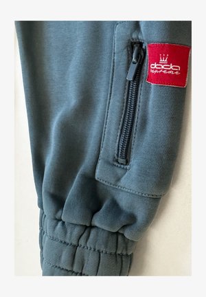 Dark blue jogger pants made from soft fabric, featuring a zip pocket and a red logo patch with a crown design on the side.