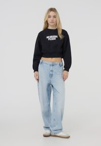 Sudadera nera crop con il testo bianco "NO DRAMA PLEASE"; abbinata a jeans in denim a vita alta di colore azzurro chiaro; calzature casual.