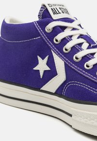 Fialové plátěné vysoké tenisky s bílými akcenty, které mají hvězdové logo, bílé tkaničky a černou gumovou podrážku s texturovanými detaily.