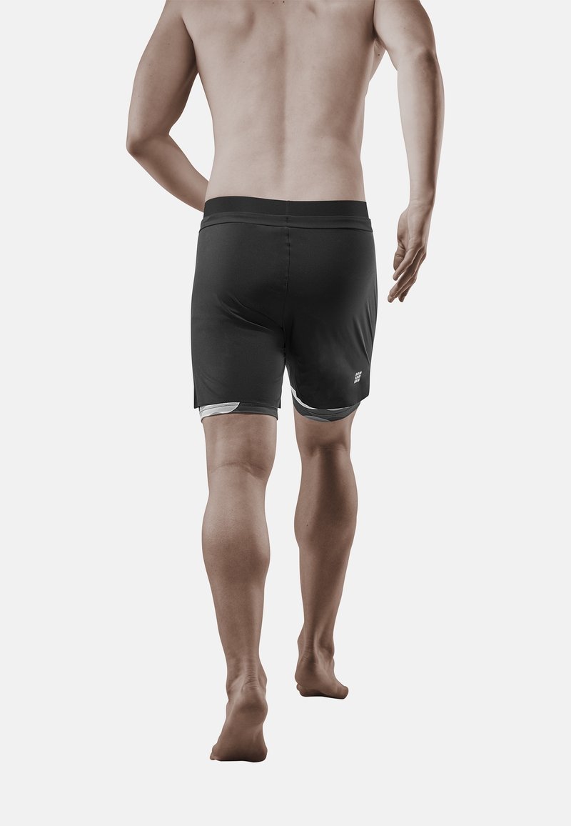 Homme portant un short de sport noir superposé, vu de dos, marchant pieds nus sur un fond blanc uni.