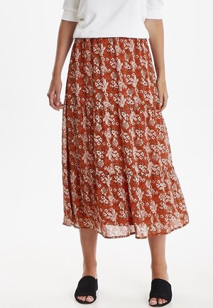 Femme portant une jupe midi couleur rouille avec un motif floral blanc, associée à un haut blanc et des chaussures noires slip-on à bout ouvert.