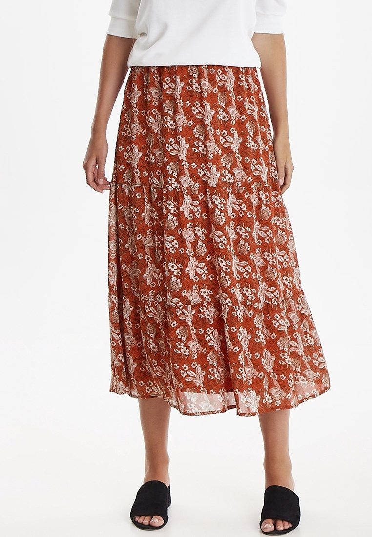Femme portant une jupe midi couleur rouille avec un motif floral blanc, associée à un haut blanc et des chaussures noires slip-on à bout ouvert.