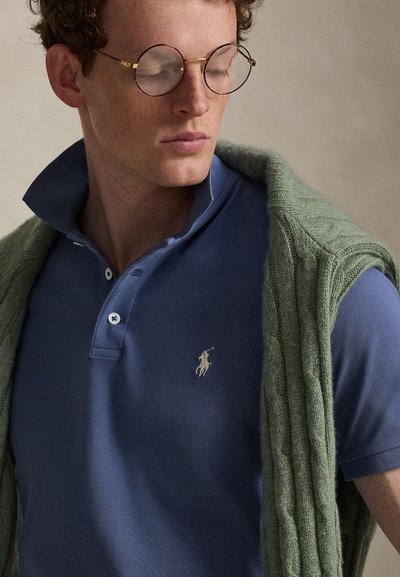 Jeune homme aux cheveux bouclés et aux lunettes rondes, portant un polo bleu et un pull vert en maille torsadée posé sur ses épaules.