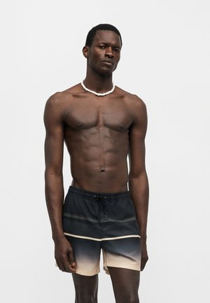 Homme torse nu portant un short de bain dégradé du noir au beige et un collier en perles blanches, debout devant un fond clair uni.