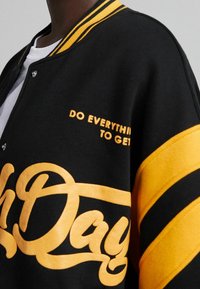 Svart varsityjacka med gula ränder och text. Har tryckknappar, mjuk textur och stor stiliserad skriptlogotyp på framsidan.