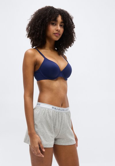 Γυναίκα με σγουρά μαλλιά που φοράει μπλε μπρά και γκρι σορτς Polo Ralph Lauren, στέκεται μπροστά σε απλό ανοιχτό φόντο.