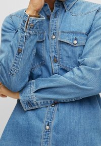 Camisa de mezclilla azul claro con frente de botones, dos bolsillos en el pecho y mangas largas. Presenta un cuello puntiagudo y detalles de botones a presión.