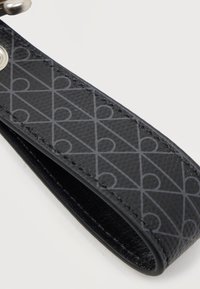 Μαύρο λουράκι από υφή δέρματος με γεωμετρικό διαγώνιο μοτίβο καρδιάς και μεταλλικό κρίκο στην επάνω αριστερή γωνία.
