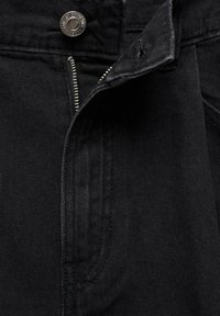 Mustat farkut, joissa on hopeinen metallipainike ja vetoketjuhytti, näkyvät saumat ja teksturoitu pinta.