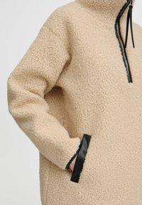 Beige fleece pullover med høj krave, med sorte læderdetaljer og sidelommer. Det strukturerede stof giver varme og komfort.