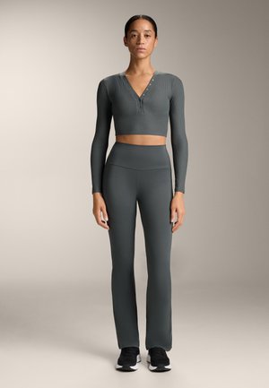 Sivi rebrasti dugih rukava crop top s V-izrezom i detaljem s gumbima, uparen s visokim sivim tajicama. Crne atletske cipele.