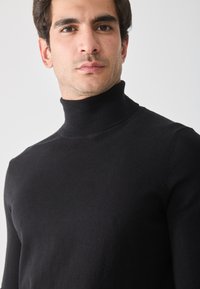 Suéter negro de cuello alto hecho de tejido de punto suave. Presenta un diseño ajustado con un cuello alto y mangas largas. Detalles de textura menores visibles.