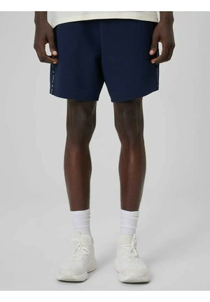 Mężczyzna w granatowych sportowych szortach, białych skarpetkach do kostek i białych sportowych butach, stojący na tle jednolitego jasnoszarego tła.