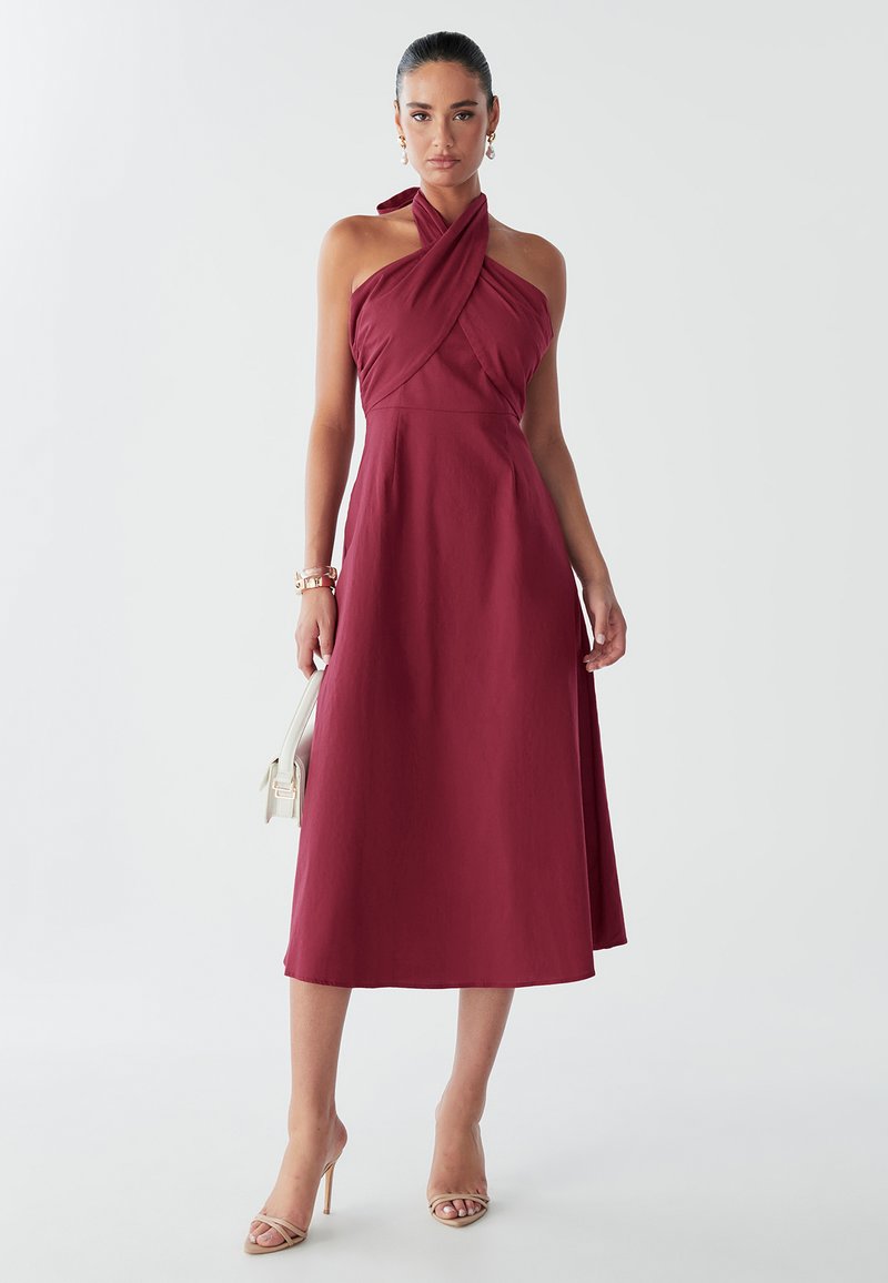 Robe bordeaux à col licou avec un corsage ajusté et une jupe évasée, présentant une texture lisse. Le mannequin tient un petit sac blanc et porte des talons couleur nude.