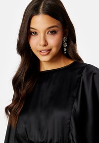 Černé saténové šaty s kulatým výstřihem a nabíranými rukávy. Modelka má na sobě třpytivé visící náušnice. Jemná textura, elegantní drapérie.