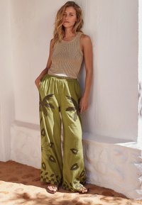 Lysebrun ribbet tanktop kombineret med løse olivengrønne bukser med mørke blomsterprint. Model poserer foran en hvid væg.