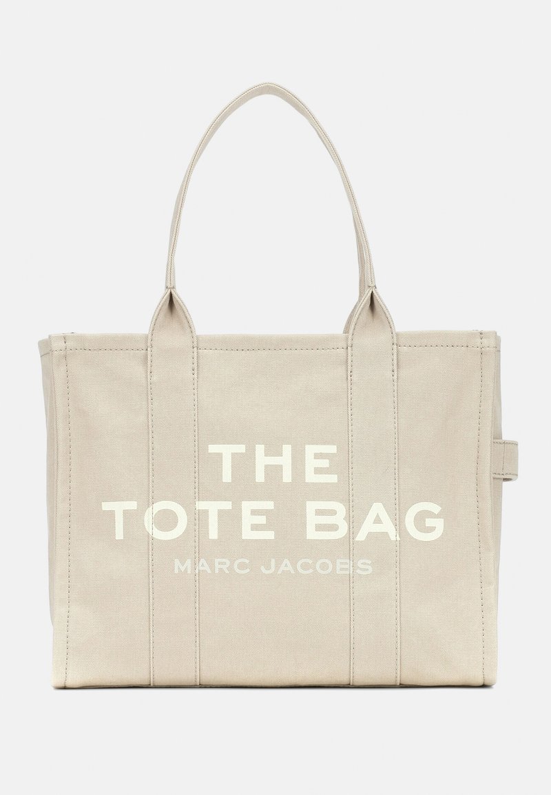 Grande bolsa tote bege feita de tela, apresentando costuras verticais, duas alças e texto impresso "THE TOTE BAG MARC JACOBS" em branco.