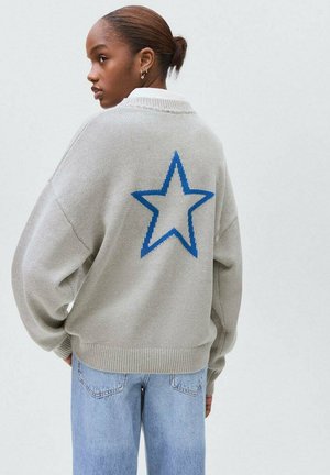 Jeune femme avec les cheveux en chignon, portant un pull léger gris avec une grande étoile bleue dans le dos et un jeans bleu clair, debout de profil.