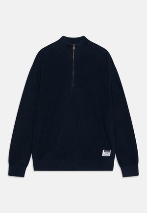 Pull en maille côtelée marine avec col haut et fermeture éclair mi-hauteur, avec un petit logo cousu en bas à gauche. Texture douce.