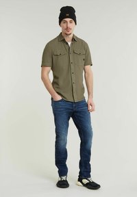 Korte mouwen olijfgroen shirt met twee borstzakken, gecombineerd met blauwe jeans en zwarte sneakers. De persoon draagt ook een zwarte beanie.