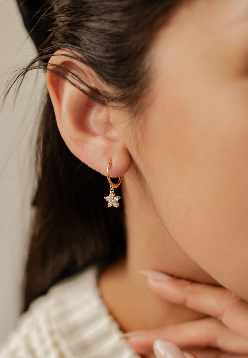 Pendiente de aro dorado con un charm de estrella colgante, adornado con strass transparentes. La oreja y la mano son parcialmente visibles, destacando el pendiente.