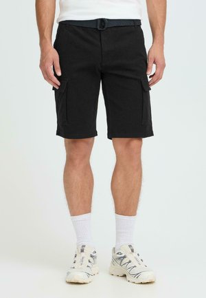 Mand iført sorte cargo-shorts, der går til knæene med sidelommer, hvide sokker og hvide sportssko, stående mod en ensartet lys baggrund.