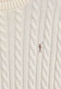 Crème kabelgebreide trui met dikke textuur ribbels en een kleine bruin geborduurde figuuraanpassing op de voorkant.