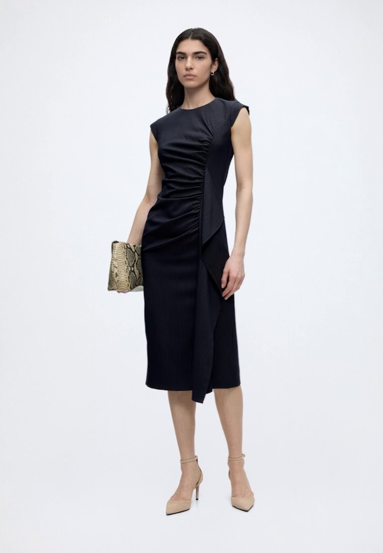 Femme portant une robe midi noire sans manches froncée, tenant une pochette en cuir de serpent et portant des talons béiges à bride à la cheville, debout sur fond blanc.