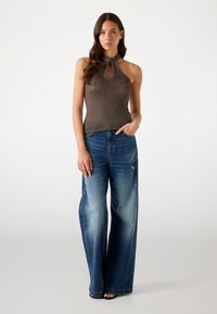 Hnědý žebrovaný halter top s detaily v oblasti klíče, spárovaný s širokými bledými modrými džíny s odřenými akcenty. Modelka stojí s jednou rukou v kapse.