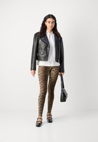 Črna usnjena motoristična jakna, bela bluza, legice s leopardjim vzorcem in čevlji črne barve. Drži črno torbico. Enobarvna bela podlaga.