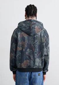 Hooded sweatshirt featuring a camouflage pattern with leaves and branches in earthy tones. Black ribbed cuffs and hem, soft texture.