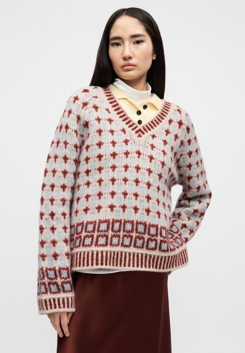 Strickpullover mit V-Ausschnitt in Hellblau, mit roten geometrischen Mustern und gerippten Akzenten. Weiche Textur, oversized Passform und lange Ärmel.