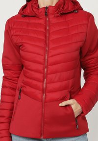 Mujer con una chaqueta acolchada roja con capucha y cremallera frontal, una mano en un bolsillo lateral con cremallera, combinada con jeans de corte claro.