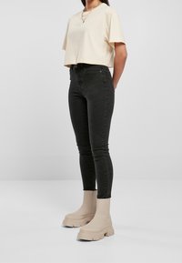 Donna con una maglietta beige corta, jeans skinny neri a vita alta e stivaletti chunky beige, in piedi contro uno sfondo neutro.