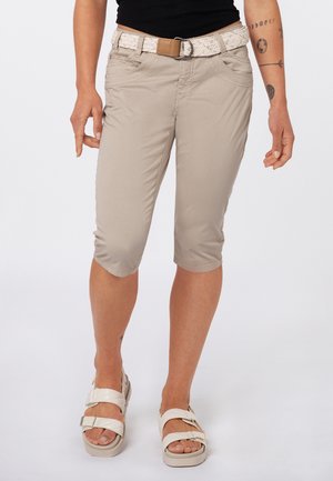 Frau trägt beige kurze Hosen mit einem geflochtenen Gürtel, schwarzes Top, weiße Sandalen und sichtbare Arm-Tattoos, steht vor einem weißen Hintergrund.
