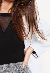 Svart topp med transparent insats, ljusgrå blazer och strukturerad grå kjol. Långt brunt hår delvis synligt. Fokusera på tyg- och designdetaljer.