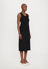 Schwarzes Tanktop mit gewelltem Saum, kombiniert mit einem schwarzen knielangen Rock. Weiße Absatzmules vervollständigen das Outfit. Glatte Textur insgesamt.