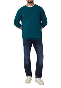 Tropfenaquafarbener Rundhalspullover mit langen Ärmeln, kombiniert mit dunklen Jeans und weißen Sneakers. Er hat eine entspannte Passform und gerippte Bündchen.