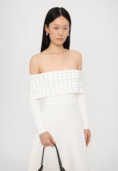 Witte off-shoulder top met studded accenten, golvende randen en lange mouwen. Het model houdt een zwarte handtas vast, gecombineerd met een witte rok.