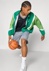 Žalias ir baltas vėjai atsparus striukė, juodas Nike marškinėliai, pilkos šortai ir balti sportbačiai. Žaidėjas dribla oranžinį krepšinį.