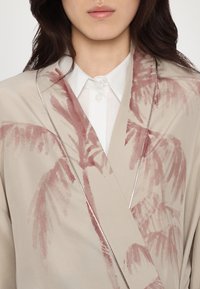 Šviesiai smėlinis kimono su rožinėmis delno lapų atspaudais, minkšta tekstūra ir sidabrinė detalė aplink iškirpę, dėvimas ant baltos palaidinės.