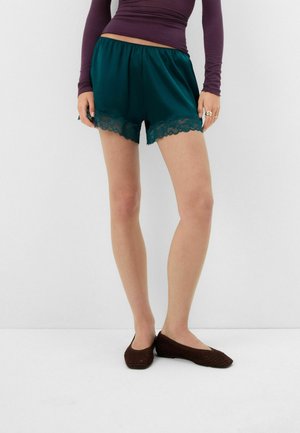 Mujer con pantalones cortos de satén verde oscuro con ribete de encaje, una camiseta de manga larga morada y zapatos planos marrones tejidos, de pie sobre un fondo blanco.